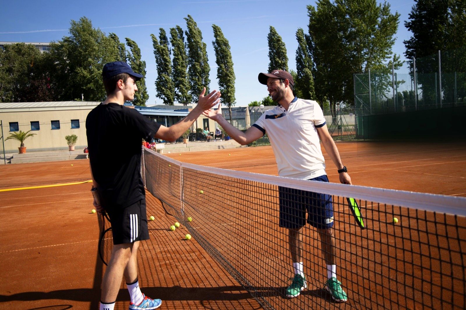 Coach und Jugendlicher klatschen sich am Netz ab, dahinter Pappeln und Sandplatz