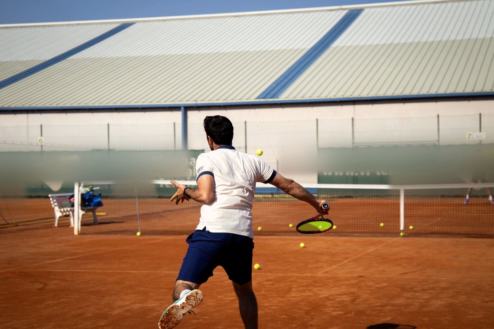 Spieler in Rückenansicht bei der Vorhand auf dem Sandplatz, im Hintergrund die Tennishalle